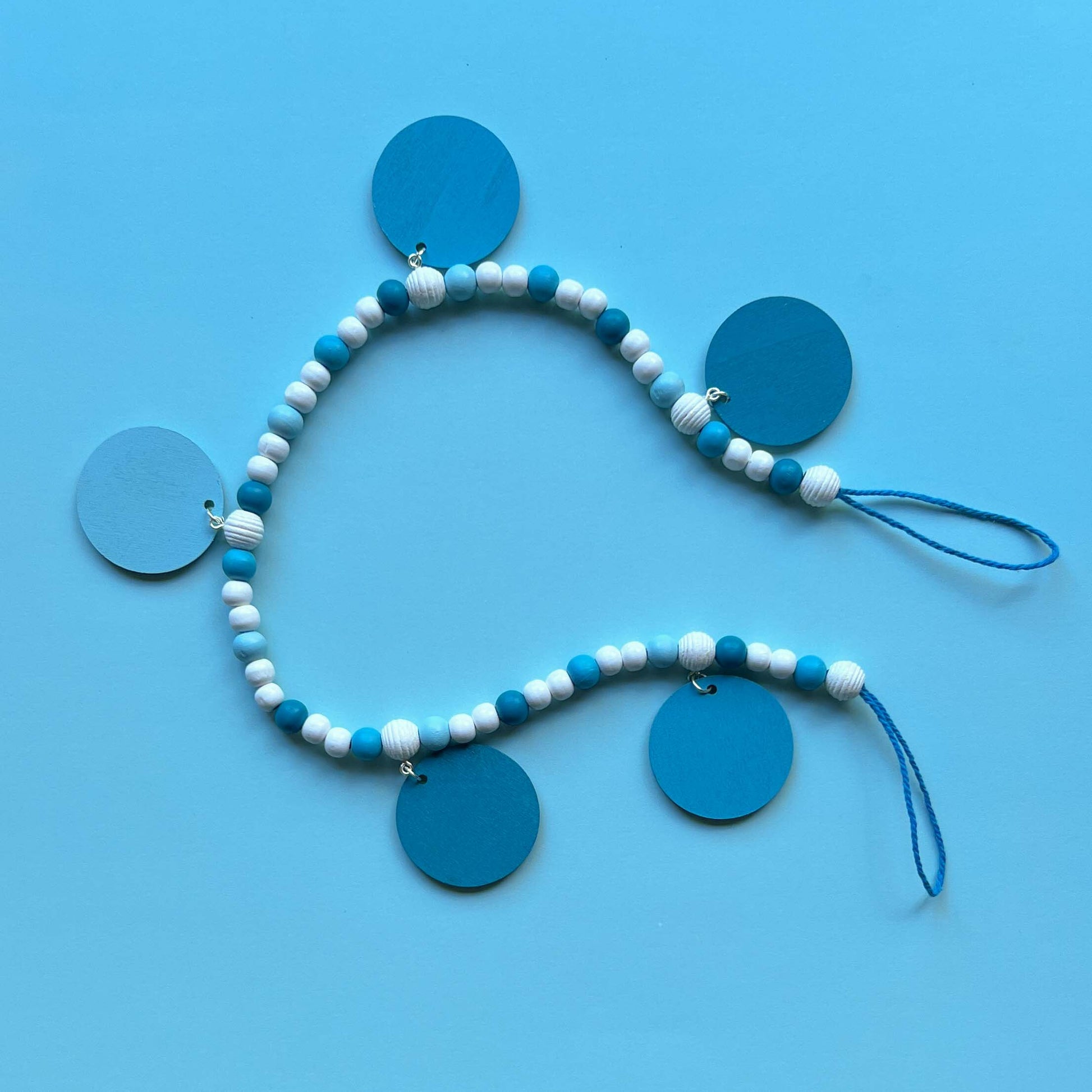 Small winter themed wood bead garland. Features white, light blue, blue, and dark blue beads with five wood circles hanging from beads. Circles include white wood snowflakes.