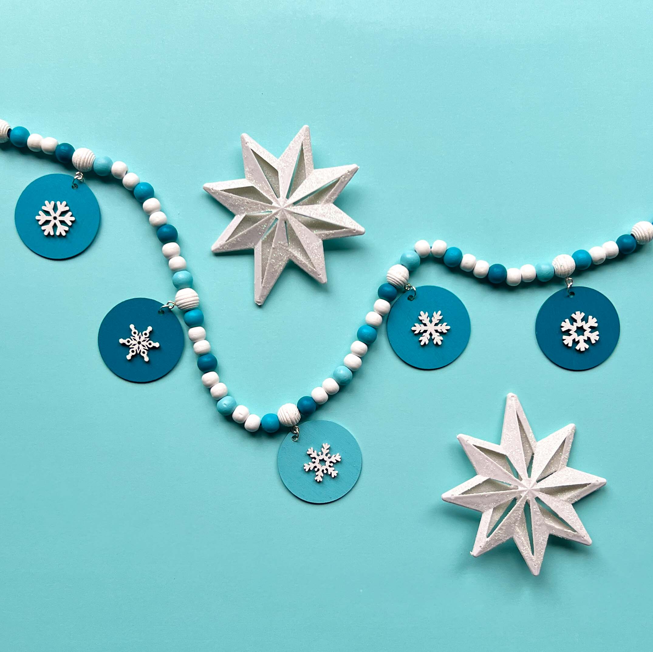 Small winter themed wood bead garland. Features white, light blue, blue, and dark blue beads with five wood circles hanging from beads. Circles include white wood snowflakes.