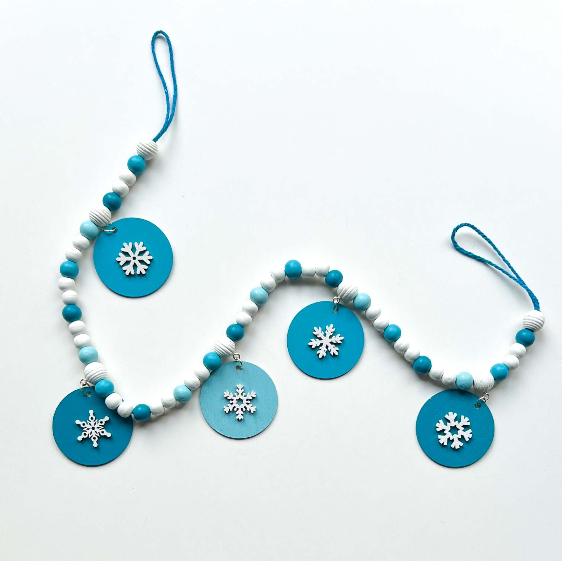 Small winter themed wood bead garland. Features white, light blue, blue, and dark blue beads with five wood circles hanging from beads. Circles include white wood snowflakes.