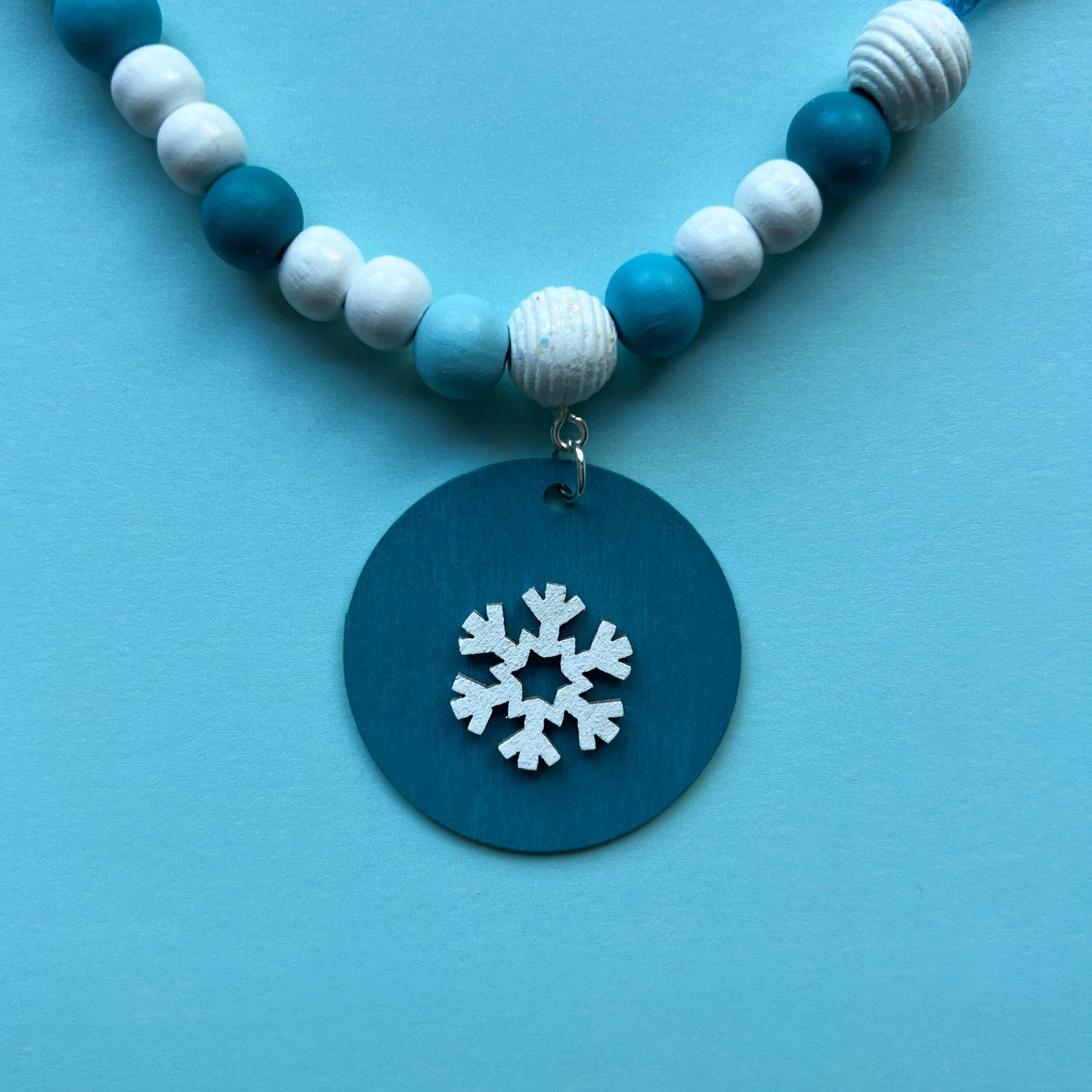 Small winter themed wood bead garland. Features white, light blue, blue, and dark blue beads with five wood circles hanging from beads. Circles include white wood snowflakes.