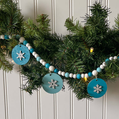 Small winter themed wood bead garland. Features white, light blue, blue, and dark blue beads with five wood circles hanging from beads. Circles include white wood snowflakes.