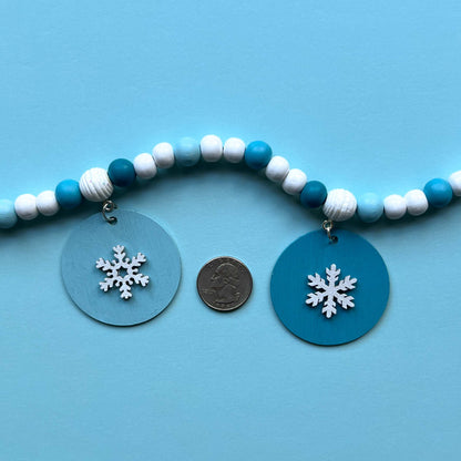 Small winter themed wood bead garland. Features white, light blue, blue, and dark blue beads with five wood circles hanging from beads. Circles include white wood snowflakes.