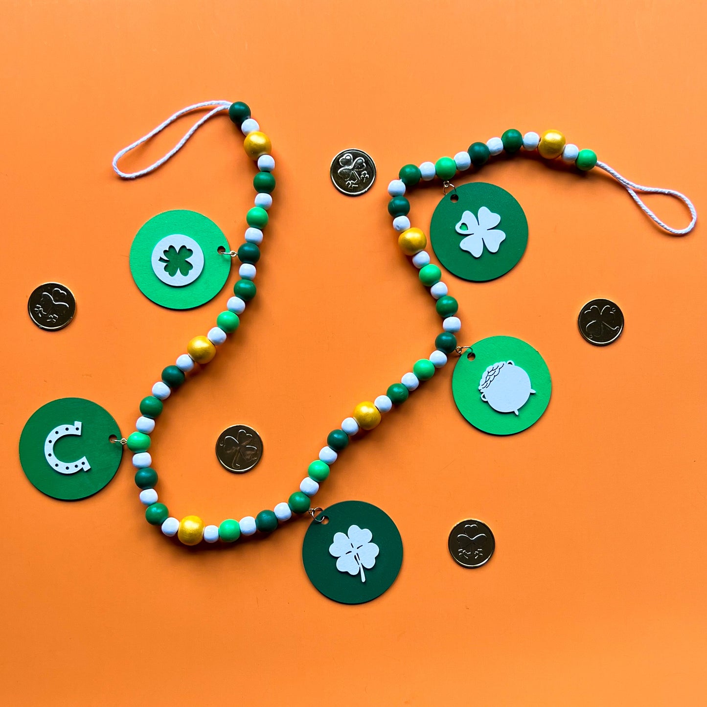 Lucky Clover Handmade Garland. The garland features five circles painted in three vibrant shades of green with white St. Patrick's Day themed icons. Circles hang from painted wood beads. St. Patrick’s Day spring seasonal decoration or gift.