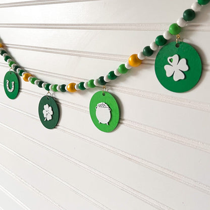 Lucky Clover Handmade Garland. The garland features five circles painted in three vibrant shades of green with white St. Patrick's Day themed icons. Circles hang from painted wood beads. St. Patrick’s Day spring seasonal decoration or gift.