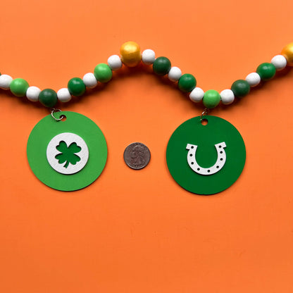 Lucky Clover Handmade Garland. The garland features five circles painted in three vibrant shades of green with white St. Patrick's Day themed icons. Circles hang from painted wood beads. St. Patrick’s Day spring seasonal decoration or gift.