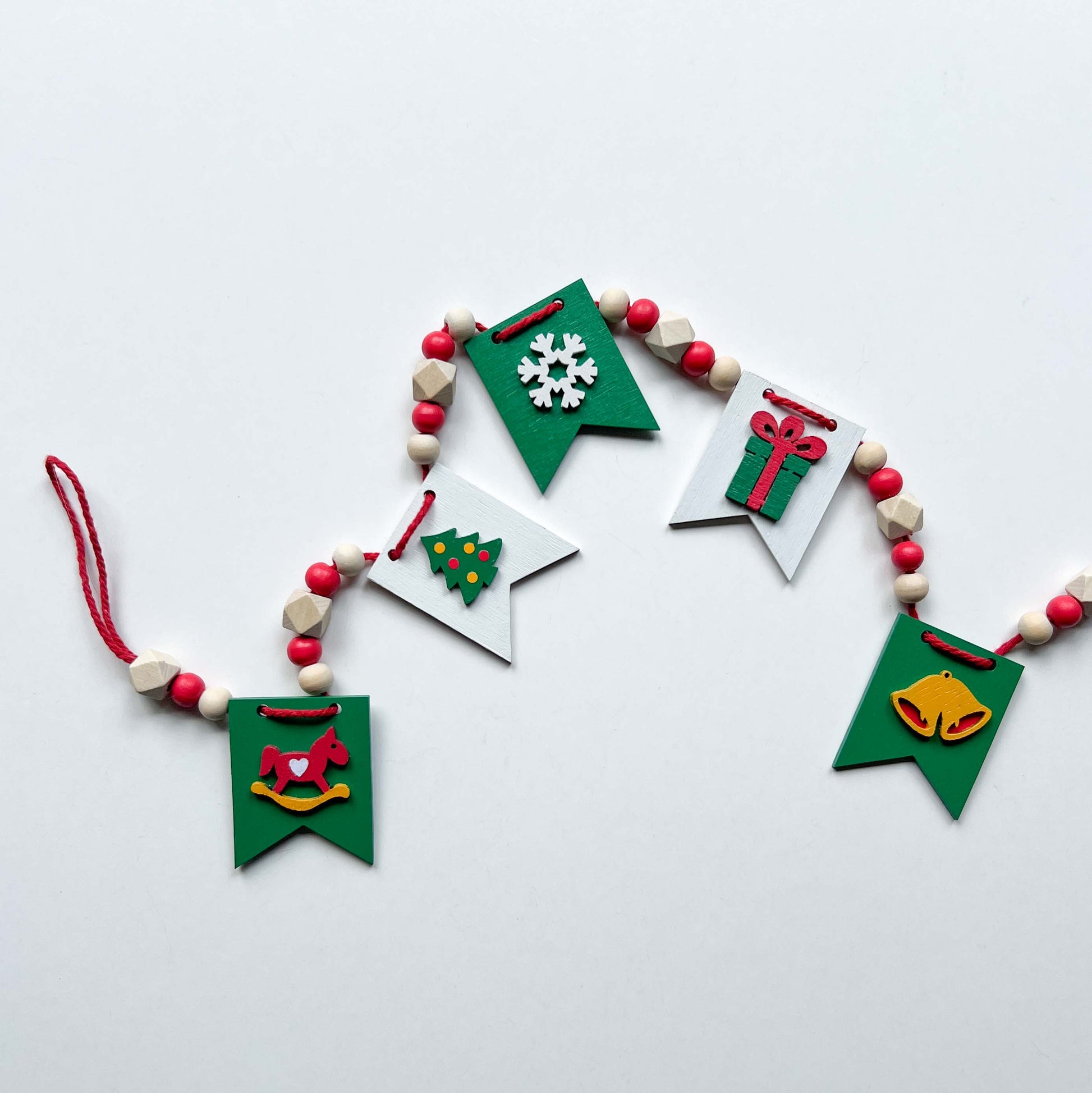 Christmas themed mini garland with pennants featuring Christmas items such as a tree, snowflake, gift, bells, and a rocking horse strung with natural and red beads.