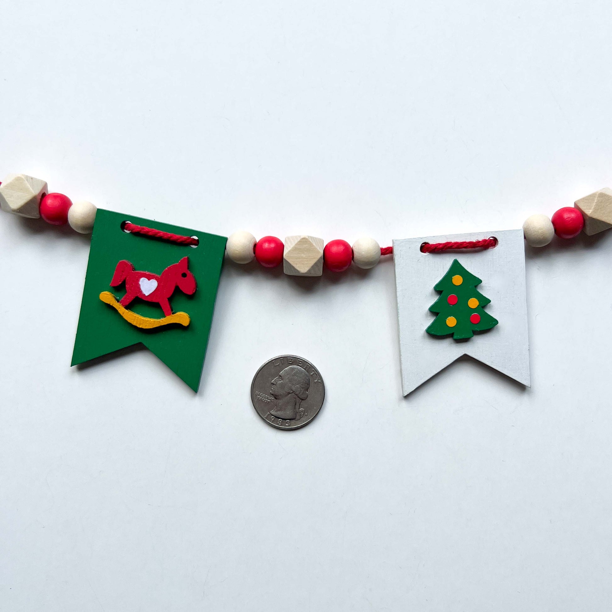 Christmas themed mini garland with pennants featuring Christmas items such as a tree, snowflake, gift, bells, and a rocking horse strung with natural and red beads.