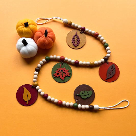 Image shows a wood bead garland with 2" wood circles that feature various colored fall leaves. Circles hang from painted beads with natural wood beads interspersed. Beads are strung on 2mm orange macrame cord with 3" loops on each end.