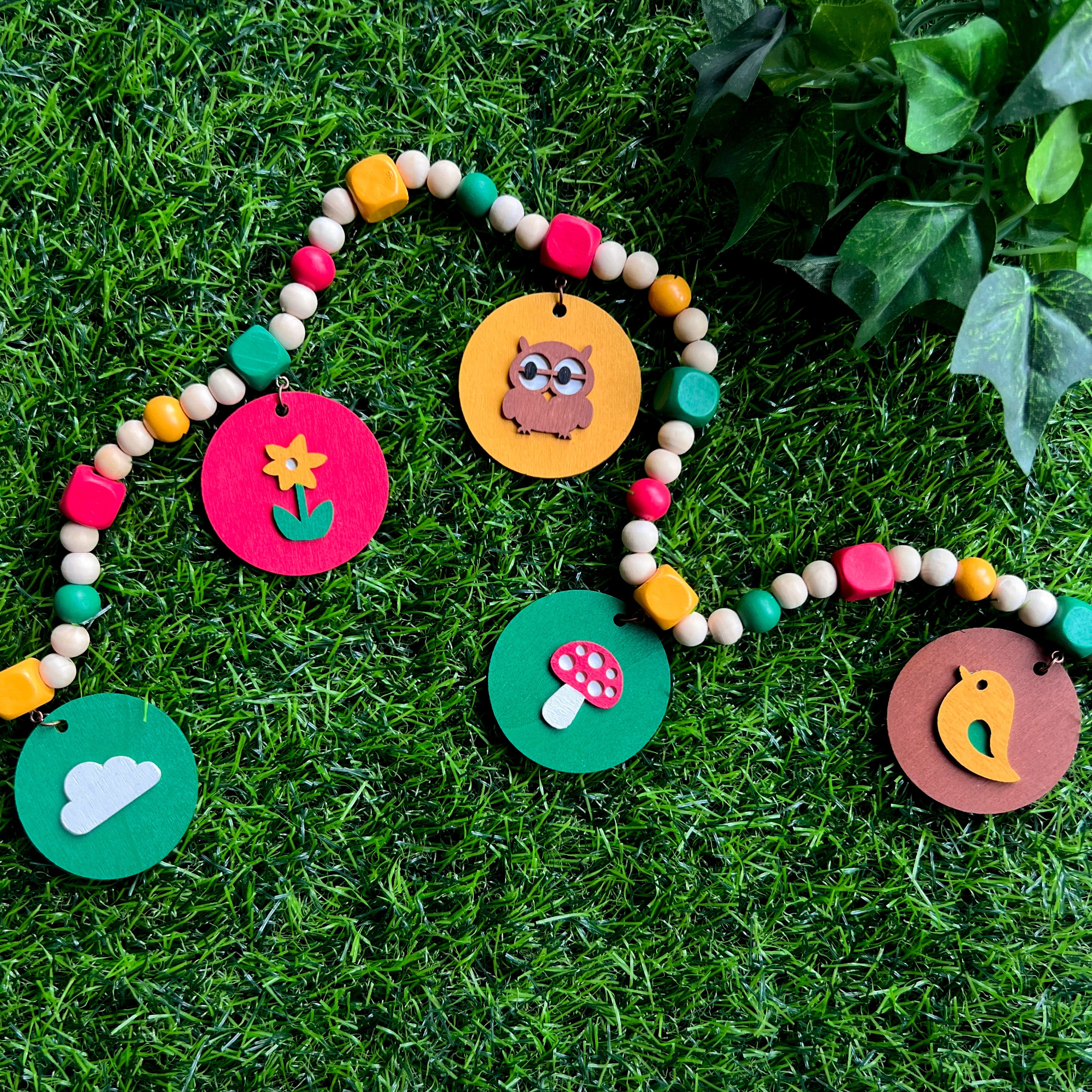 Image shows handmade garland with wood circles featuring nature themed shapes. Wood circles hang from painted square beads and are strung with other painted and natural round wood beads. Includes 3-inch loops on either end.