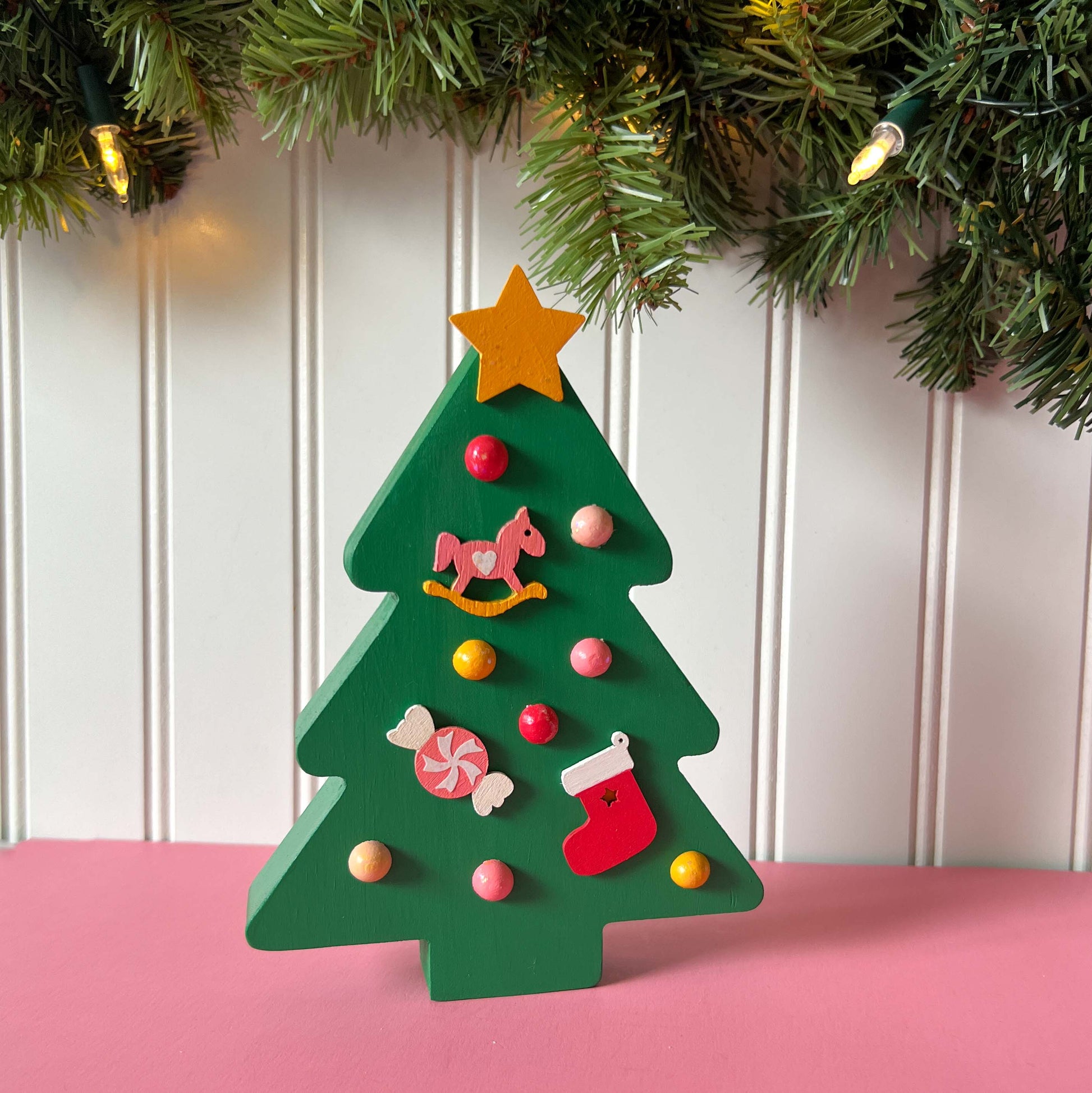 Wood Christmas tree shelf sitter painted green. Tree features a yellow star with glitter, half round wood balls painted yellow and various shades of pink. Tree also features a pink and yellow rocking horse wood ornament, a pink and white candy wood ornament, and a red and white stocking wood ornament.