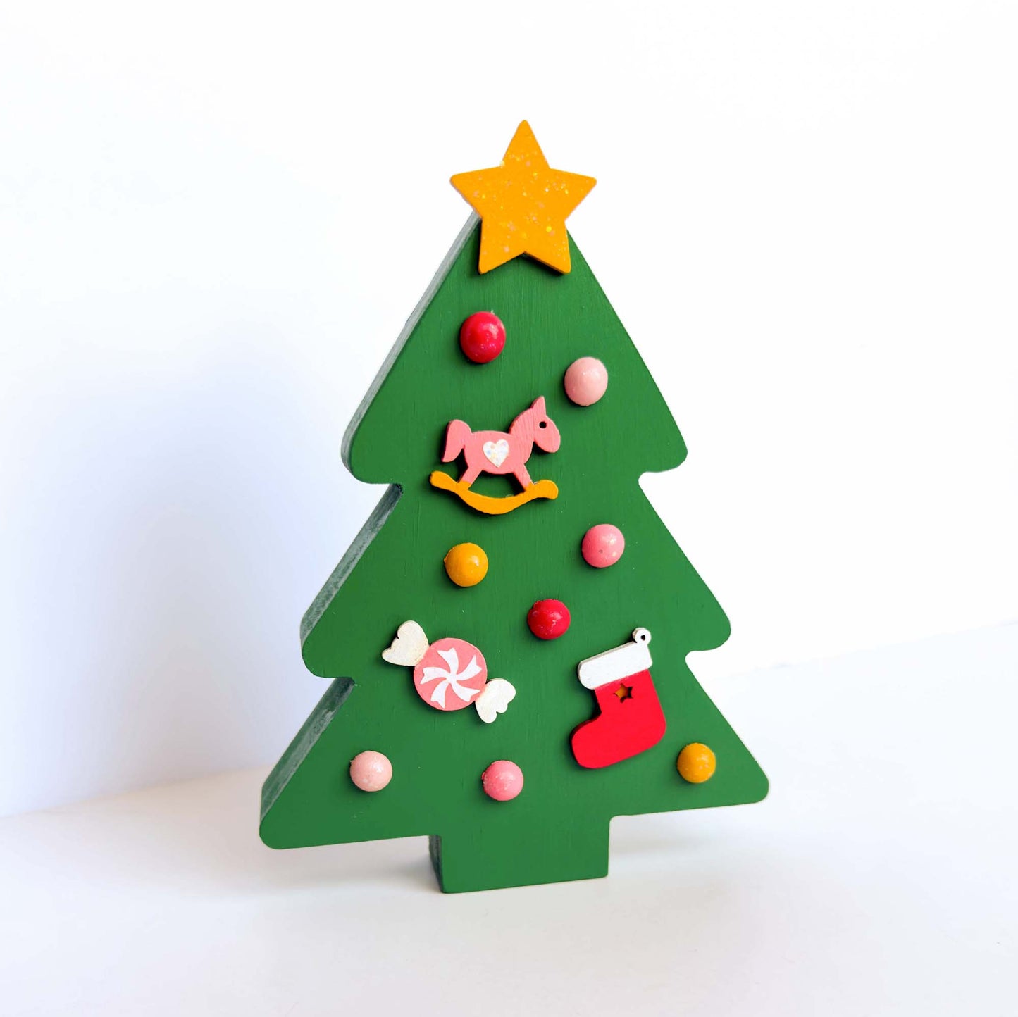 Wood Christmas tree shelf sitter painted green. Tree features a yellow star with glitter, half round wood balls painted yellow and various shades of pink. Tree also features a pink and yellow rocking horse wood ornament, a pink and white candy wood ornament, and a red and white stocking wood ornament.