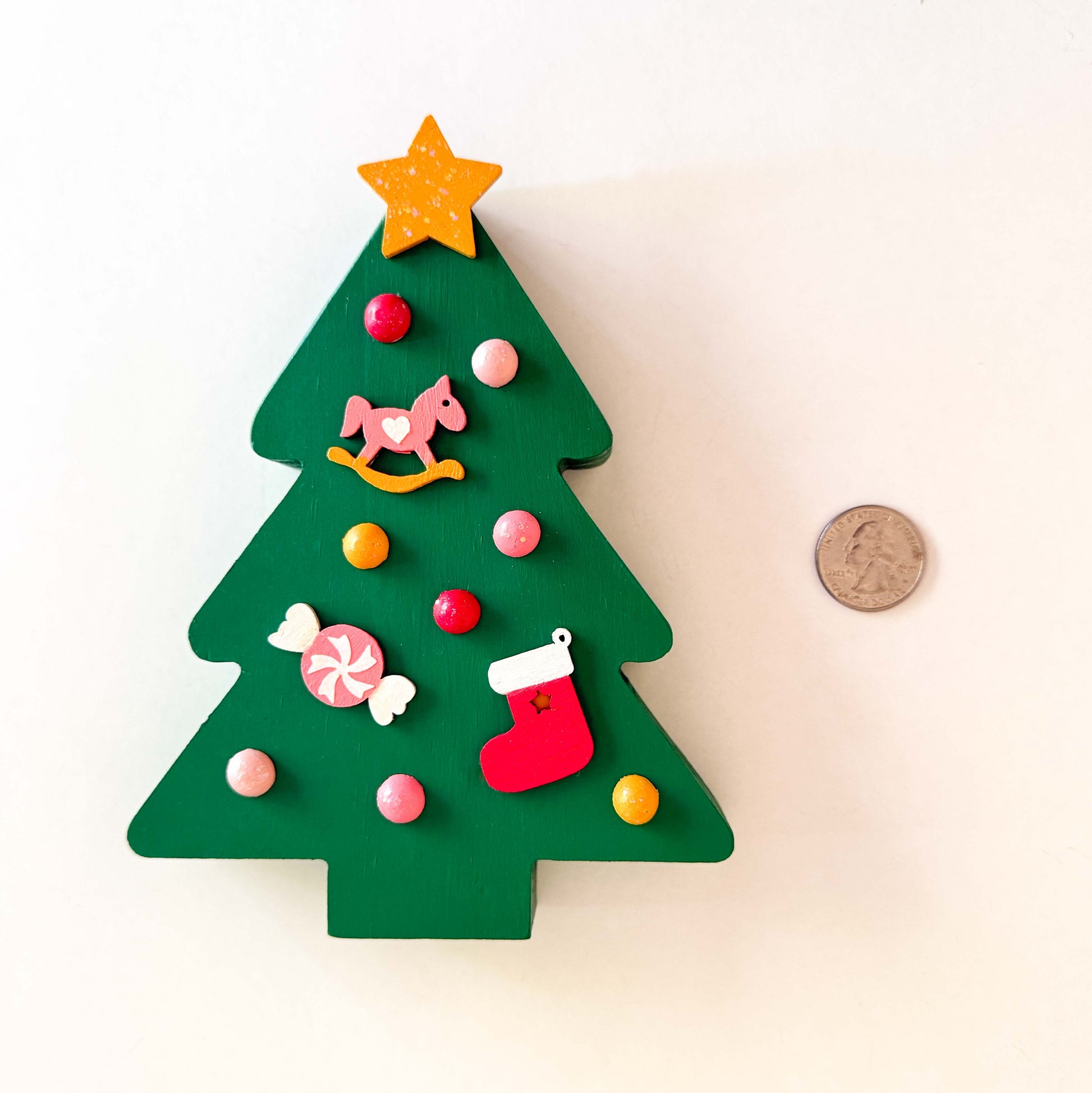Wood Christmas tree shelf sitter painted green. Tree features a yellow star with glitter, half round wood balls painted yellow and various shades of pink. Tree also features a pink and yellow rocking horse wood ornament, a pink and white candy wood ornament, and a red and white stocking wood ornament.