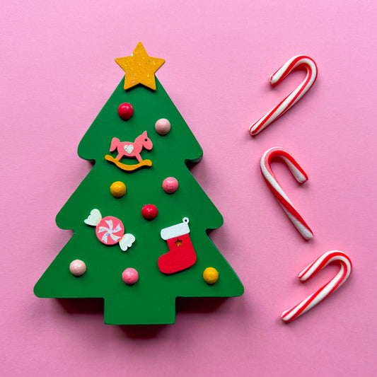 Wood Christmas tree shelf sitter painted green. Tree features a yellow star with glitter, half round wood balls painted yellow and various shades of pink. Tree also features a pink and yellow rocking horse wood ornament, a pink and white candy wood ornament, and a red and white stocking wood ornament.