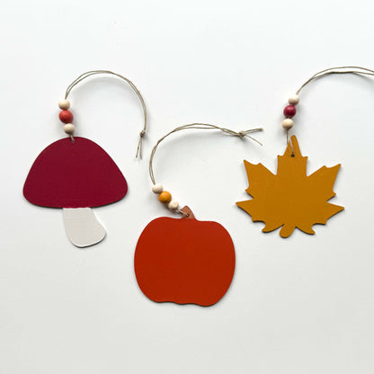 Image of the back of set of three autumn themed ornaments. The ornaments include a pumpkin, a maple leaf, and a toadstool. Each ornament hangs from twine and feature three wood beads.