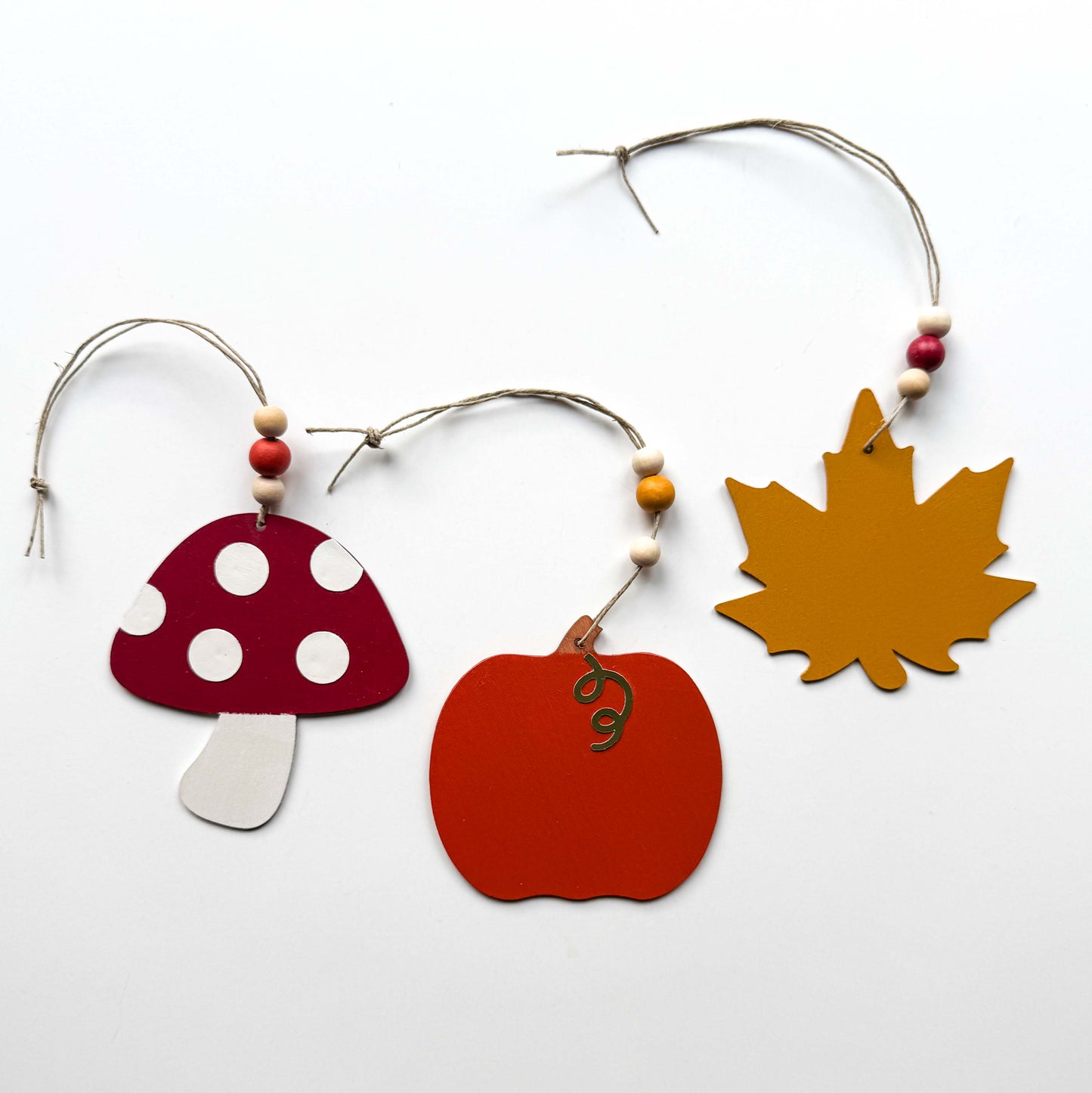 Set of three autumn themed ornaments. The ornaments include a pumpkin, a maple leaf, and a toadstool. Each ornament hangs from twine and feature three wood beads.