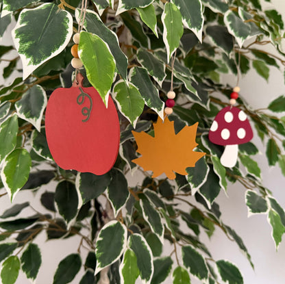 Set of three autumn themed ornaments. The ornaments include a pumpkin, a maple leaf, and a toadstool. Each ornament hangs from twine and feature three wood beads. Ornaments hang from a decorative indoor tree.