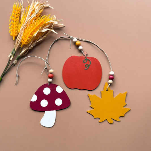 Set of three autumn themed ornaments. The ornaments include a pumpkin, a maple leaf, and a toadstool. Each ornament hangs from twine and feature three wood beads.