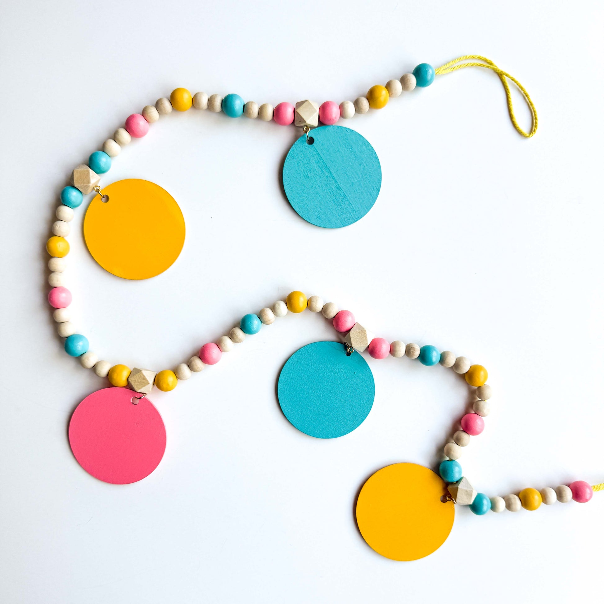 Back of summer themed wood bead garland with natural, pink, blue, and yellow beads. 5 wood circles hang from geometric beads and feature tropical themed shapes. Garland is placed on a white background.