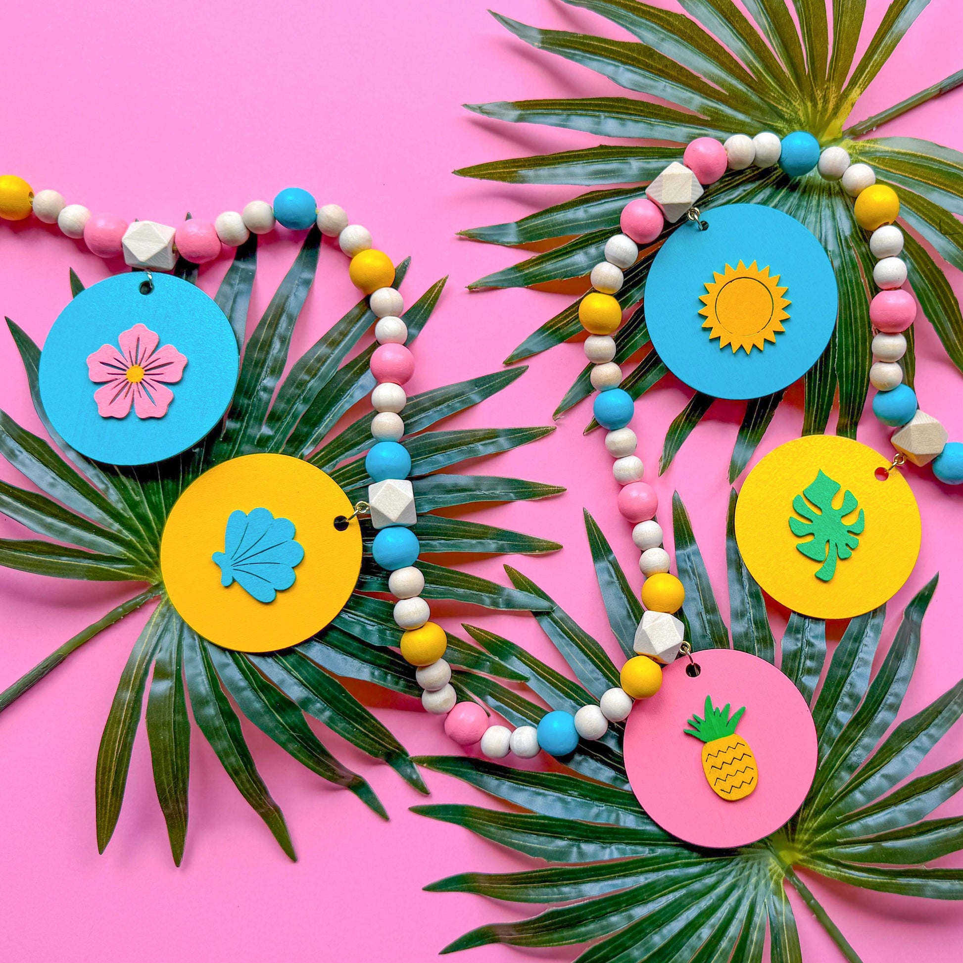 Summer themed wood bead garland with natural, pink, blue, and yellow beads. 5 wood circles hang from geometric beads and feature tropical themed shapes. Garland is placed on a pink background with palm leaves.