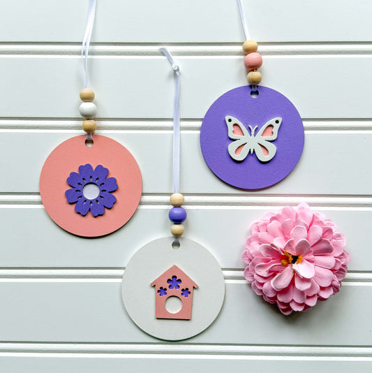 Three spring themed ornaments in white, lavender, and light pink. Ornaments each feature a different spring shape - a flower, a birdhouse, or a butterfly. Each ornament hangs from a white ribbon with three wood beads as an accent.