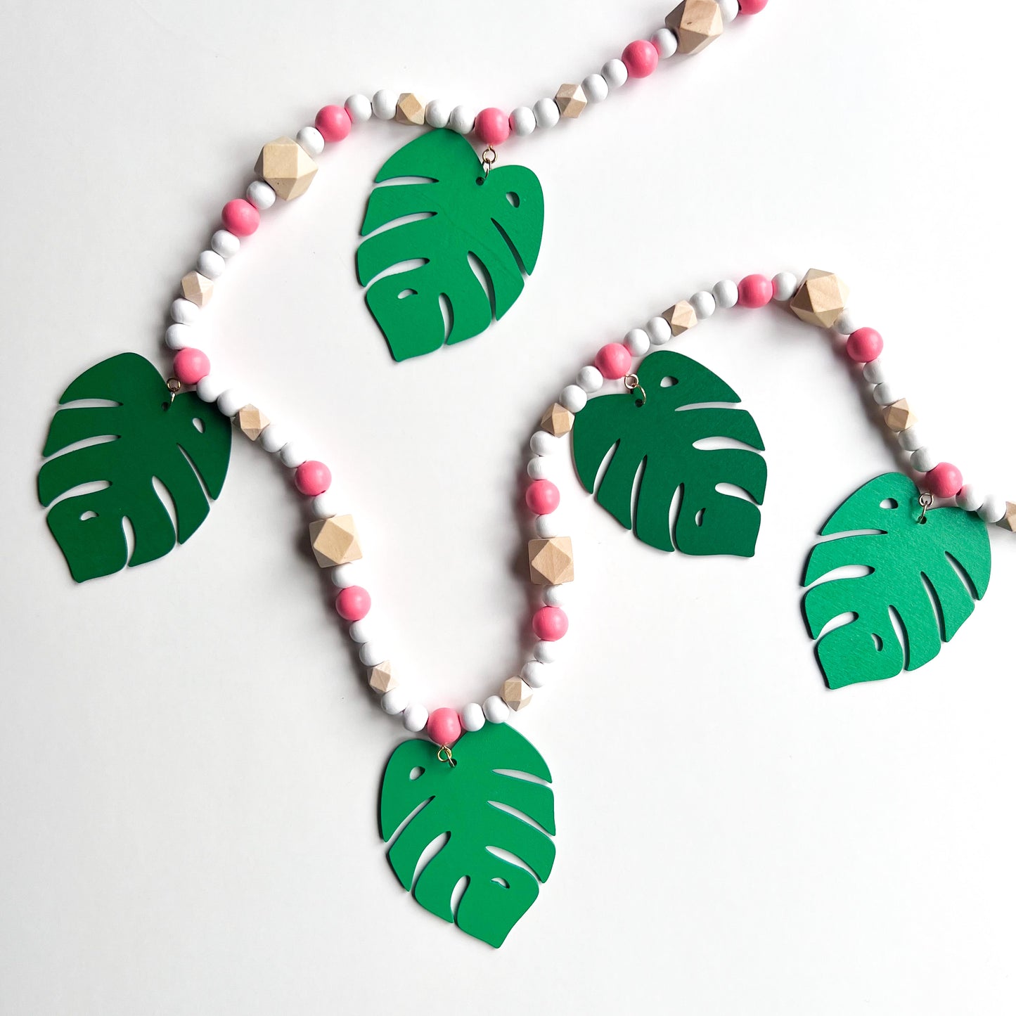 Tropical Breeze Handmade Garland. Green monstera palm leaves hang from painted pink wood beads. Garland includes natural geometric beads.