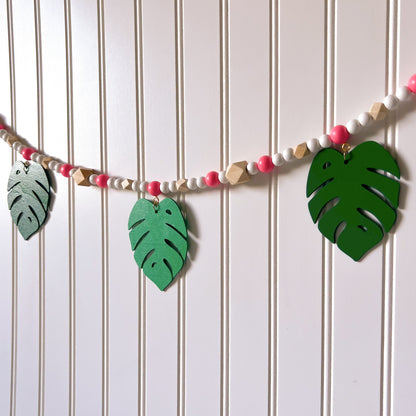 Tropical Breeze Handmade Garland. Green monstera palm leaves hang from painted pink wood beads. Garland includes natural geometric beads.