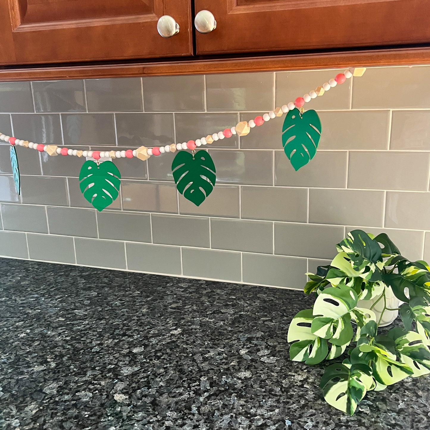 Tropical Breeze Handmade Garland. Green monstera palm leaves hang from painted pink wood beads. Garland includes natural geometric beads.