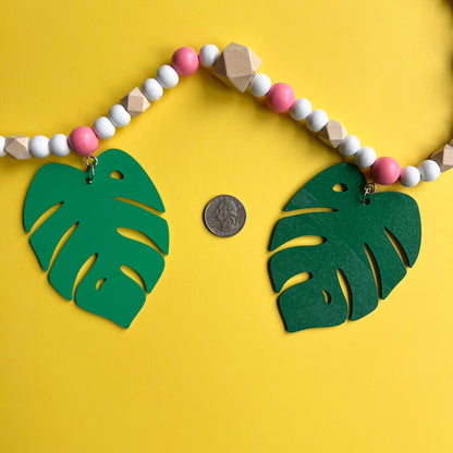 Tropical Breeze Handmade Garland. Green monstera palm leaves hang from painted pink wood beads. Garland includes natural geometric beads.