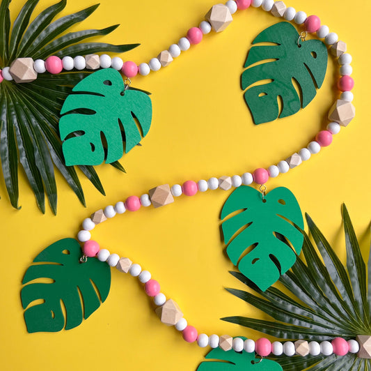 Tropical Breeze Handmade Garland. Green monstera palm leaves hang from painted pink wood beads. Garland includes natural geometric beads.