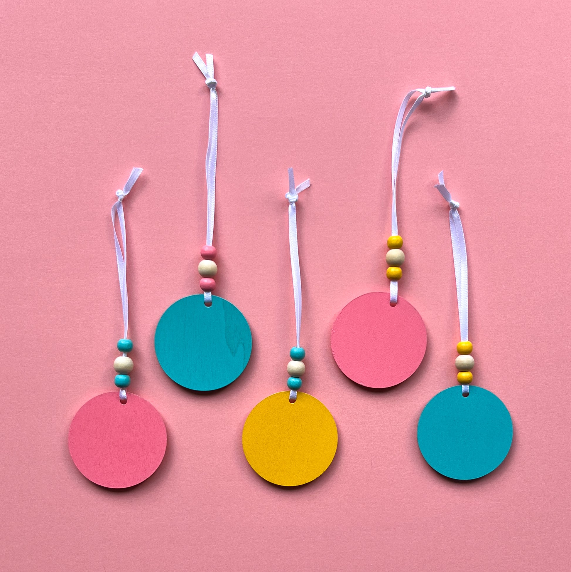 Tropical Vacay Handmade Ornament Set of 5. 2" wood circles in bright colors of pink, medium blue, and yellow feature tropical themed shapes – a flower, a drink with lemon slice, a monstera leaf, a flamingo and a pineapple.