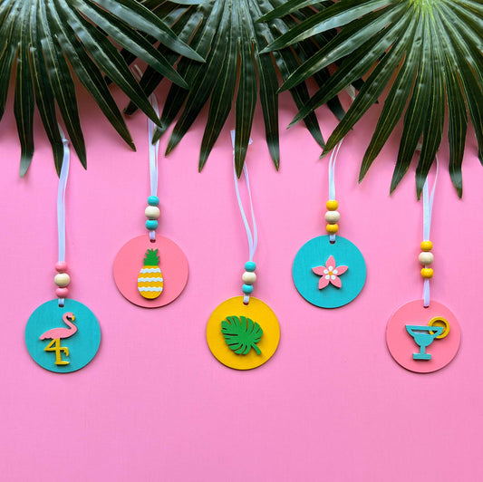 Tropical Vacay Handmade Ornament Set of 5. 2" wood circles in bright colors of pink, medium blue, and yellow feature tropical themed shapes – a flower, a drink with lemon slice, a monstera leaf, a flamingo and a pineapple.