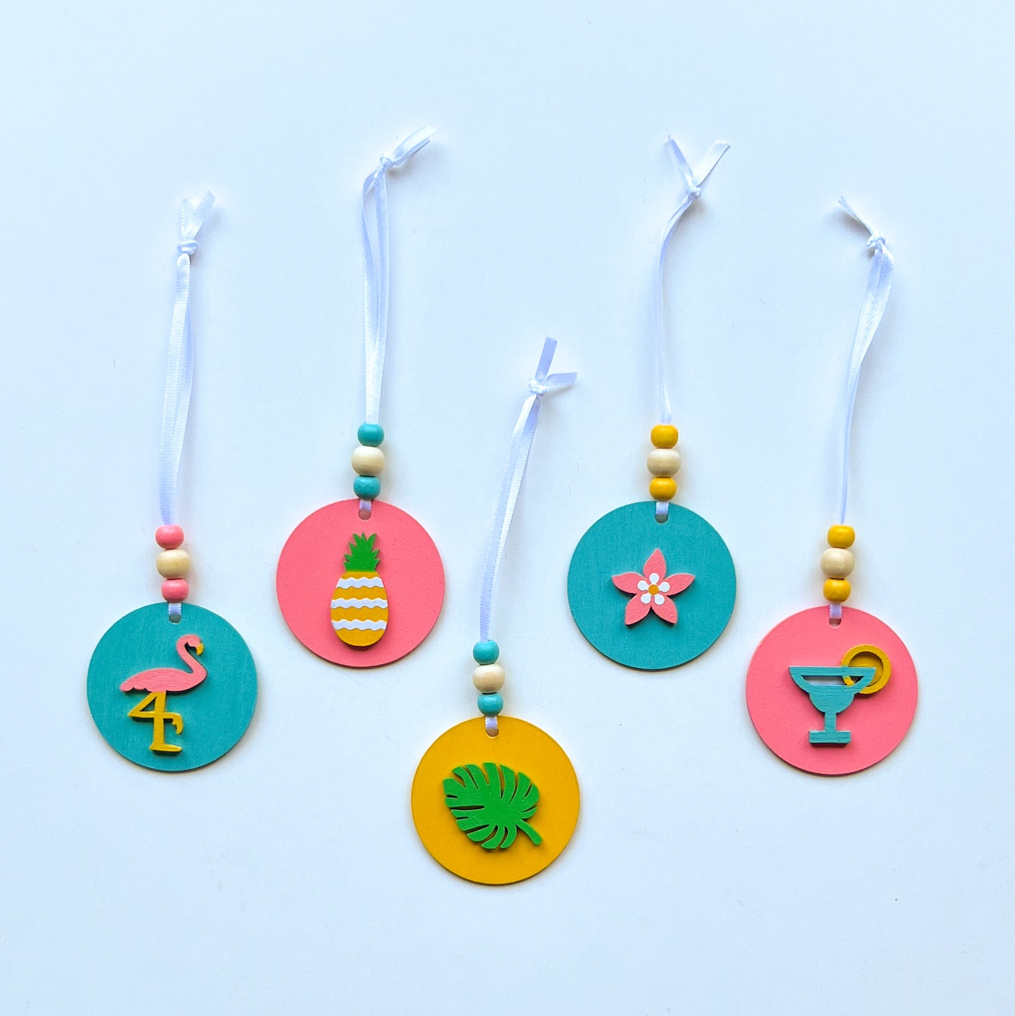 Tropical Vacay Handmade Ornament Set of 5. 2" wood circles in bright colors of pink, medium blue, and yellow feature tropical themed shapes – a flower, a drink with lemon slice, a monstera leaf, a flamingo and a pineapple.