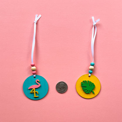 Tropical Vacay Handmade Ornament Set of 5. 2" wood circles in bright colors of pink, medium blue, and yellow feature tropical themed shapes – a flower, a drink with lemon slice, a monstera leaf, a flamingo and a pineapple.