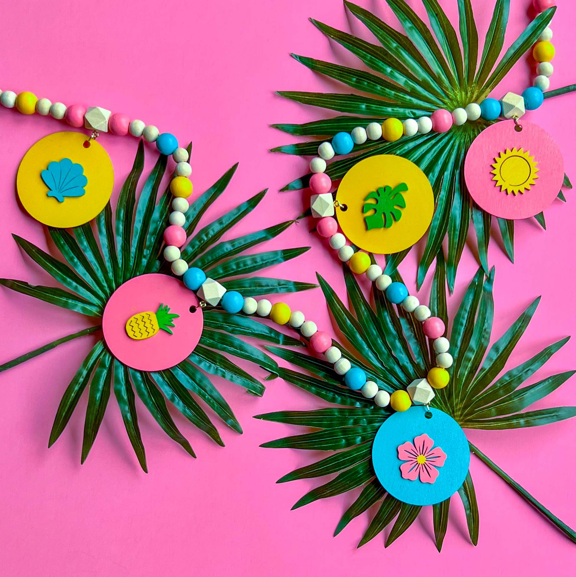 Summer Vacay Handmade Garland. The garland features wood circles with a tropical themed shape: seashells, pineapple, flower, monstera leaf, the sun. Shapes hang from wood beads. Summer tropical seasonal decoration or gift.