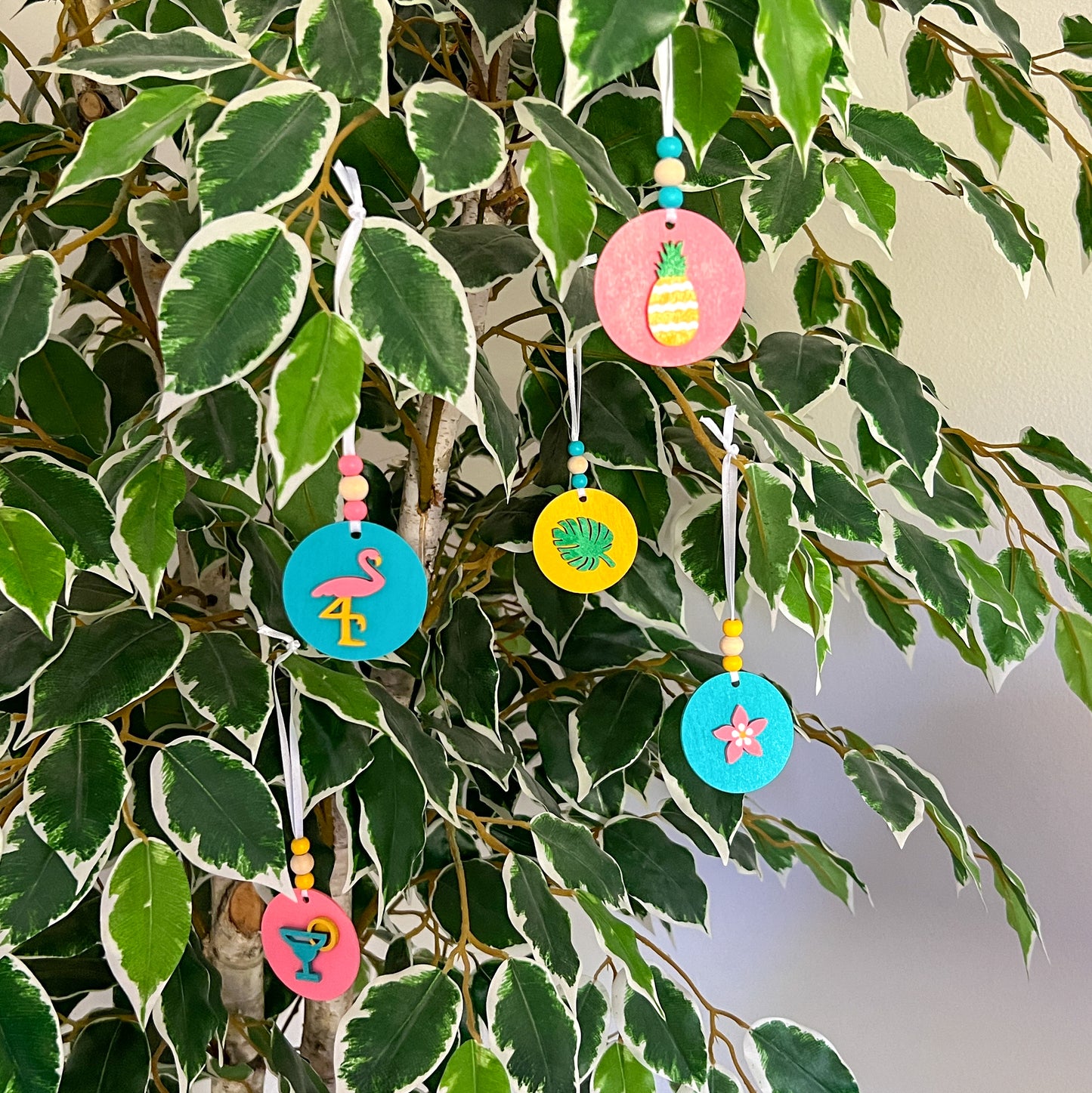 Tropical Vacay Handmade Ornament Set of 5. 2" wood circles in bright colors of pink, medium blue, and yellow feature tropical themed shapes – a flower, a drink with lemon slice, a monstera leaf, a flamingo and a pineapple.